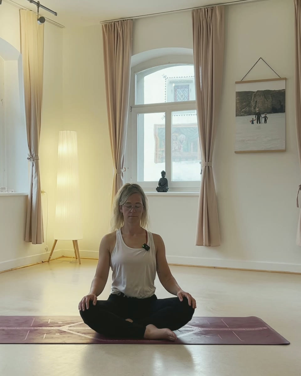 Yogalehrerin Eli sitzt mit geschlossenen Augen im Schneidersitz auf ihrer Yogamatte